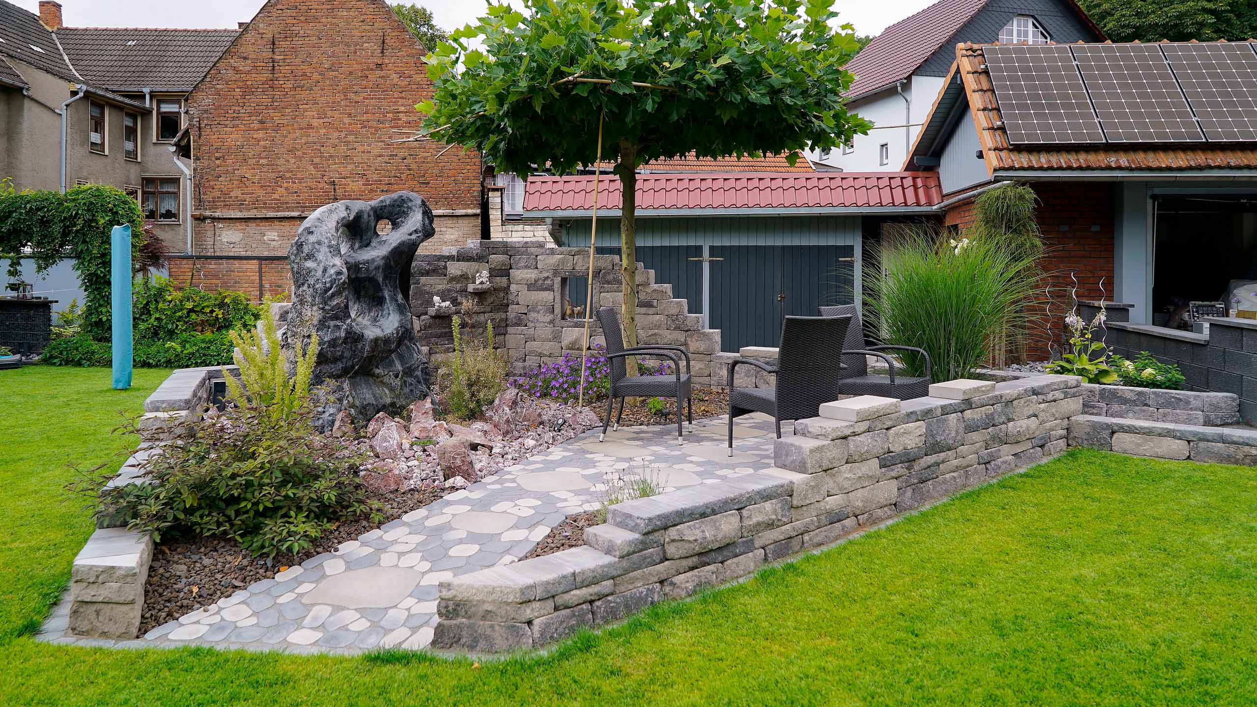 Gartengestaltung mit gepflasterter Sitzecke und Steinskulptur, erstellt von der Firma Aussengestaltung Vogt
